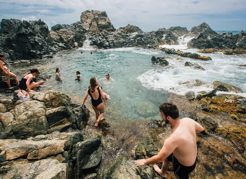 Schwimmer klettern über dunkle Felsen in den Natural Pool auf Aruba